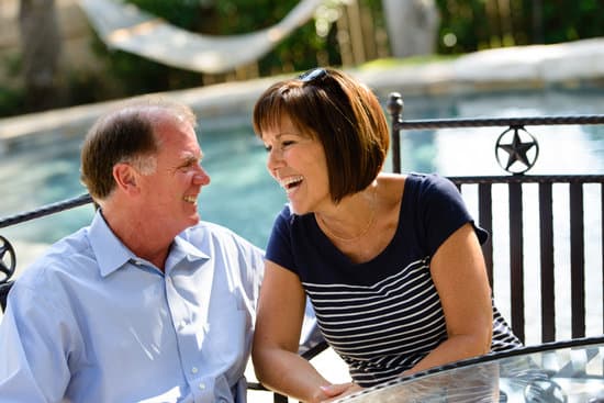 Mature adults smiling and enjoying retirement together in their backyard.  They are laughing and looking at each other lovingly.  They are sitting at a table in front of a pool with a hammock in the background.